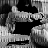 Pixelated black-and-white image of a female sitting on a bed, cradling their arms around their folded legs, surrounded by bunched-up blankets and a smartphone in the foreground.