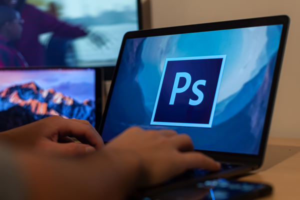 A person types on a laptop computer, the screen of which is filled with the Photoshop logo. A tablet and computer monitor are in the background to the left.