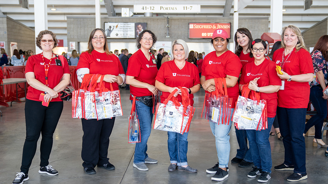 A group of eight women wearing red U Volunteer T-shirts, some holding tote bags to give away, smile for a photo in the Rice-Eccles Stadium corridor during the 2022 Employee Appreciation Day event. Image courtesy of Dave Titensor, University of Utah.