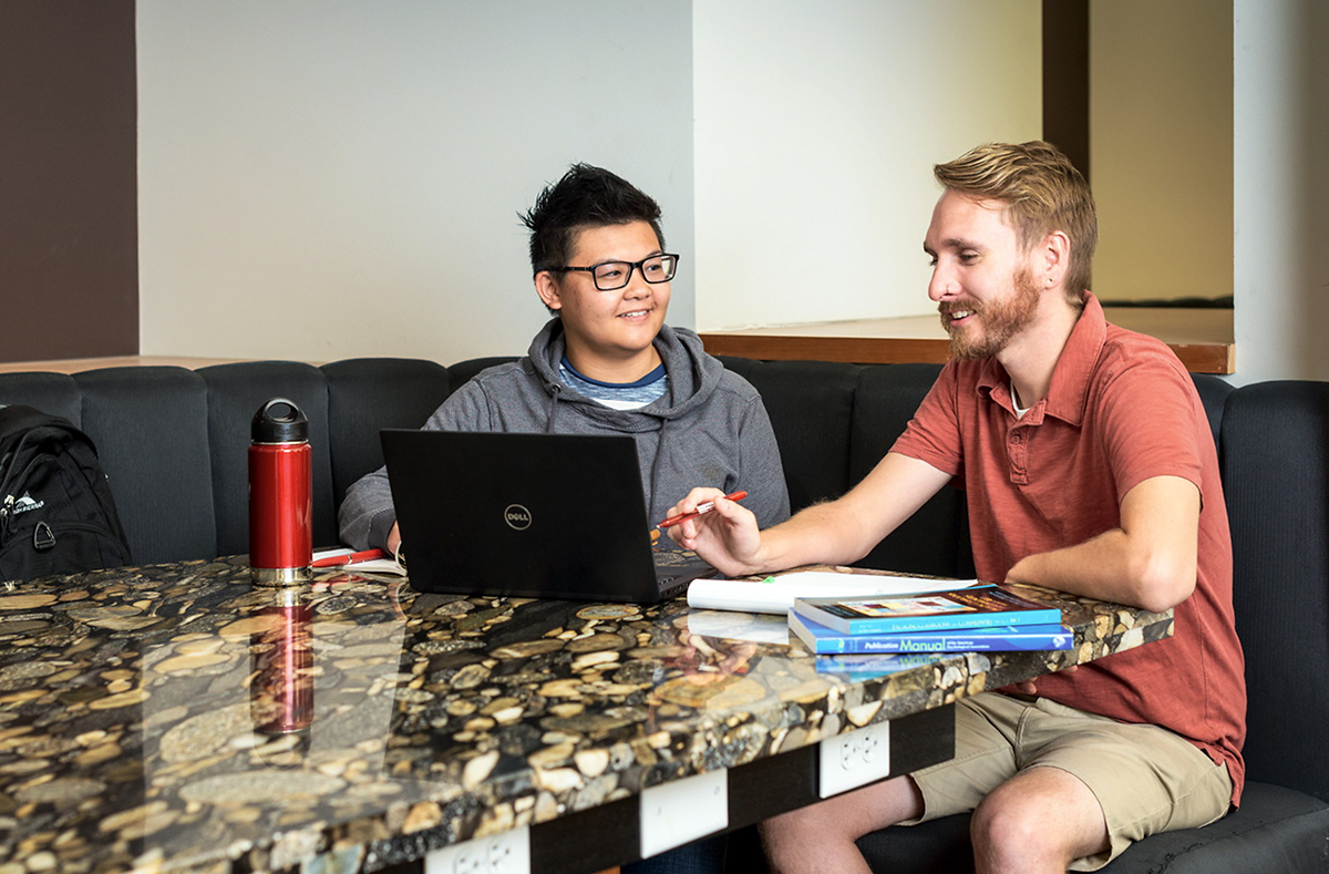 Two male University of Utah students sit at a study space with a marble countertop table in Marriott Library. The student on the left has spiky black hair, is donning glasses, and is wearing a gray hoodie. The student on the right has light brown hair and a beard, is wearing beige shorts and a salmon colored polo shirt, and is pointing at a laptop computer with a red pen in his right hand. Image courtesy of the University of Utah.