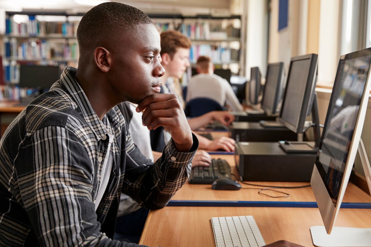 A male student in a flannel shirt with his hand brought up under his chin looks at a desktop computer’s display in a library. Other students appear in the background, along the same line of computer desks. 