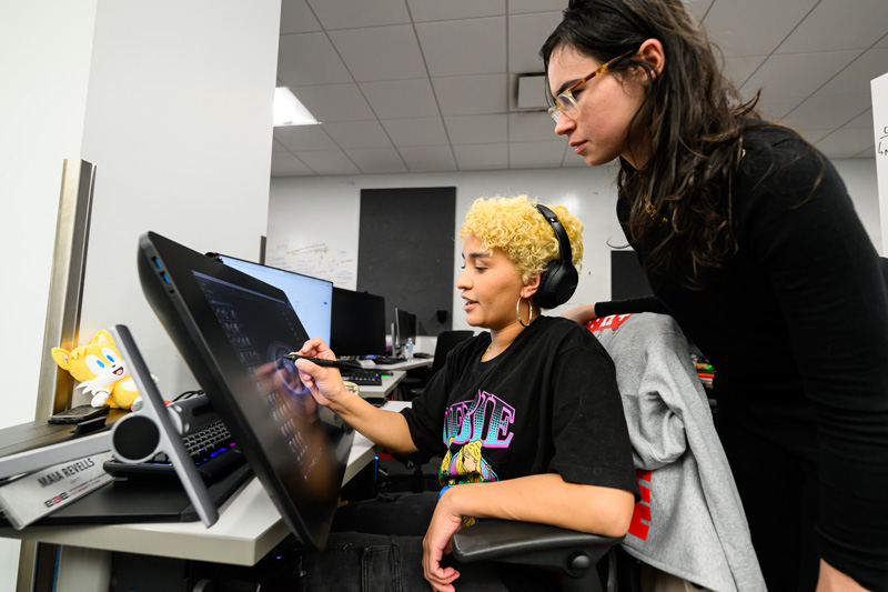 A student with curly short yellow hair and over-the-ear headphones works on a desktop computer in a University of Utah Division of Games computer lab while another student with long brown hair and glasses standing behind them watches. (Courtesy of the University of Utah)