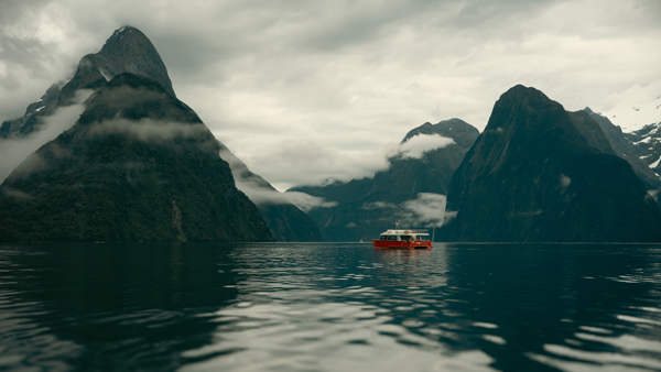 A red boat with white top sails on the waters of Milford Sound near the Southern Alps of New Zealand. Clouds hang low among the mountains in the background.