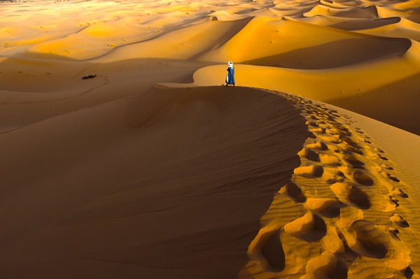 A person walks atop a dune in a golden desert.