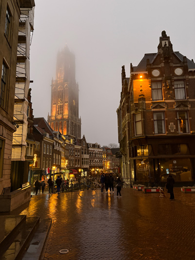 Lights glow among the gothic architecture of the Dutch city, which is shrouded in fog. About a dozen people brave the wet weather.