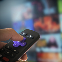 Image of a Roku streaming device remote in use with a television blurred out in the background