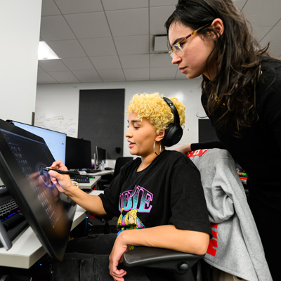 A student with curly short yellow hair and over-the-ear headphones works on a desktop computer in a University of Utah Division of Games computer lab while another student with long brown hair and glasses standing behind them watches. (Courtesy of the University of Utah)