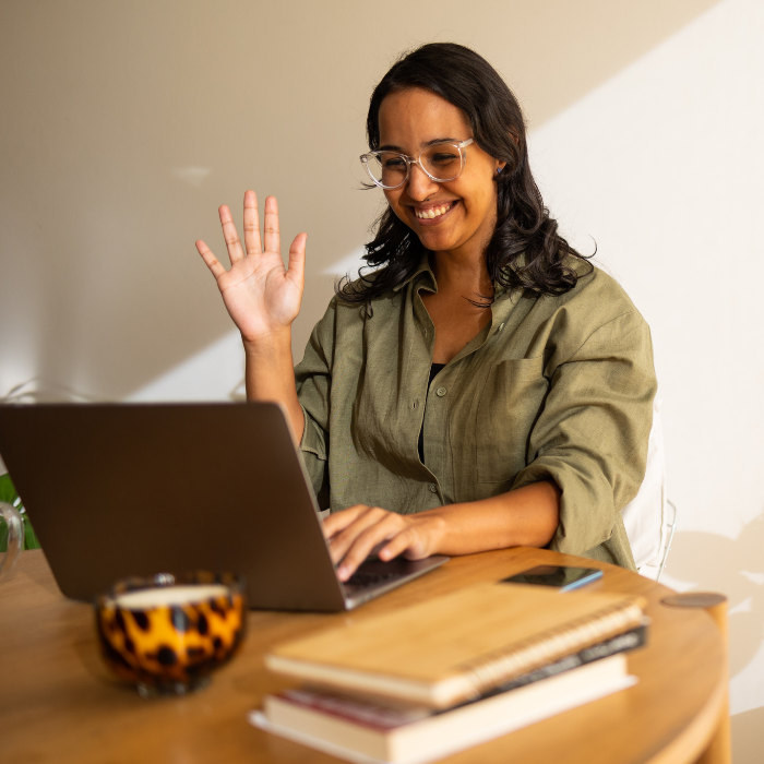 A woman sits in front of a laptop computer and waves to fellow participants in an online meeting.